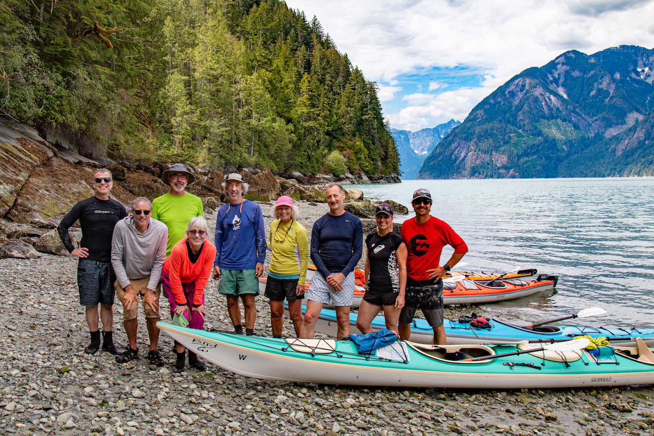 A group of kayakers posing with kayaks and mountains in the background